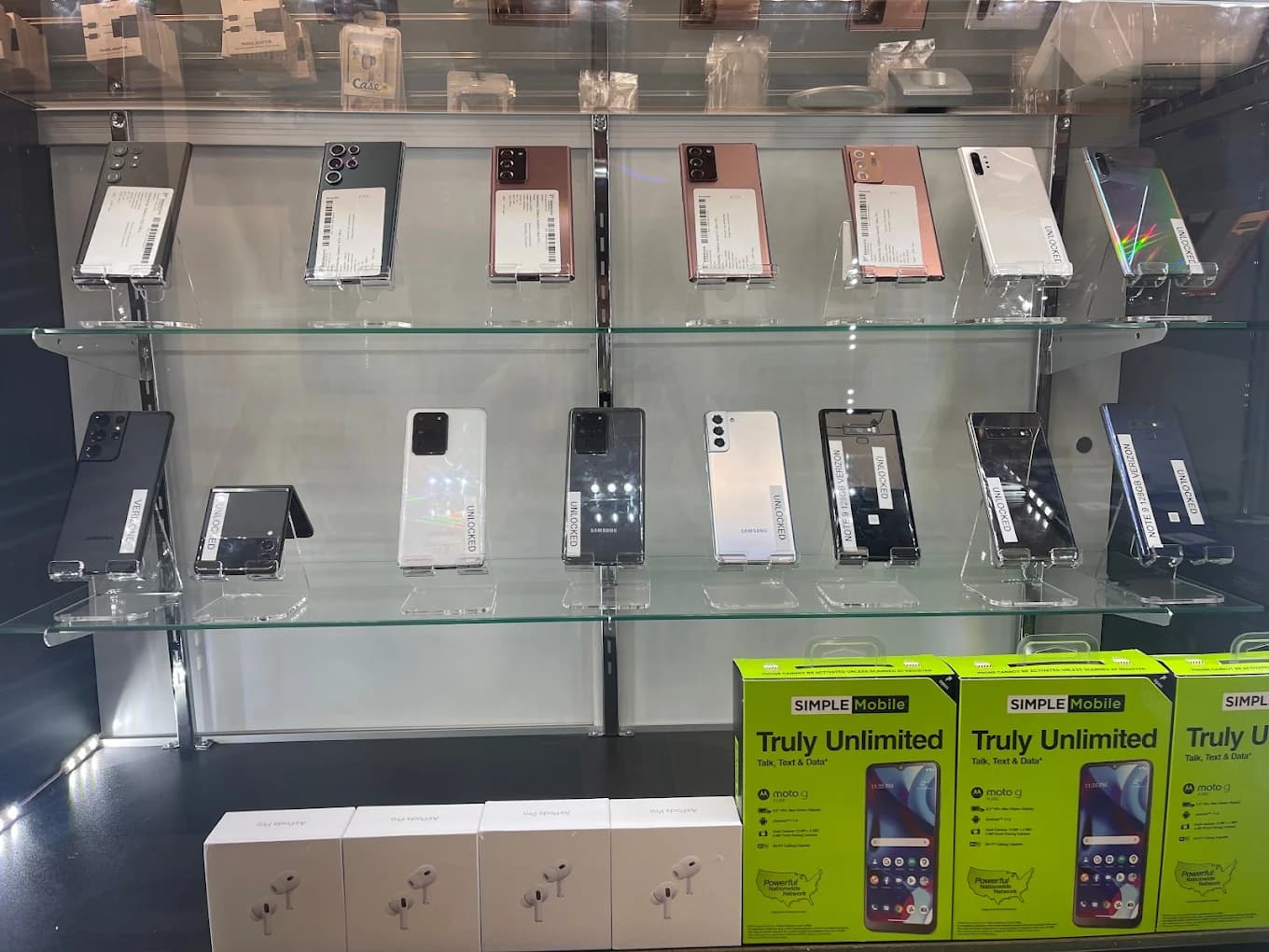 Smartphones displayed on glass shelves above boxed AirPods and Simple Mobile prepaid phone packages.