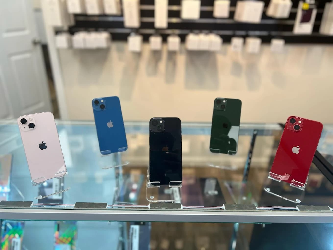 Five colorful iPhones displayed on clear stands inside a glass retail store showcase.
